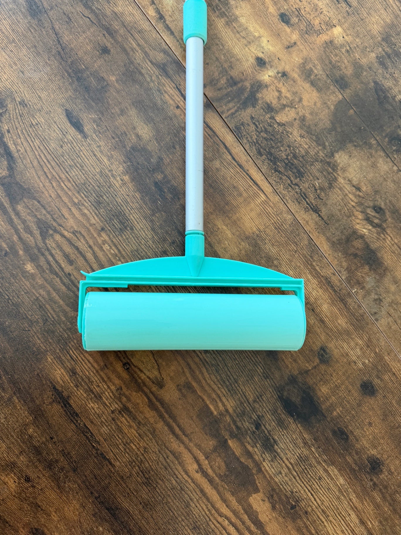 A pet hair remover stick roller with a mint green adhesive roller and handle, resting on a dark brown wooden floor.