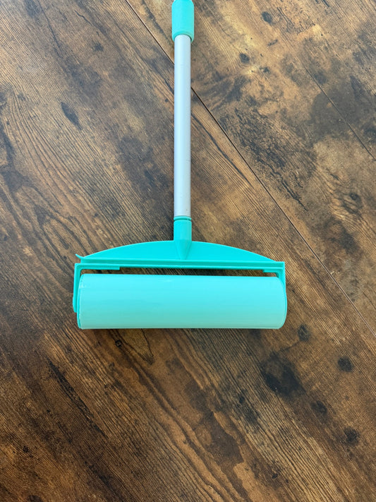 A pet hair remover stick roller with a mint green adhesive roller and handle, resting on a dark brown wooden floor.