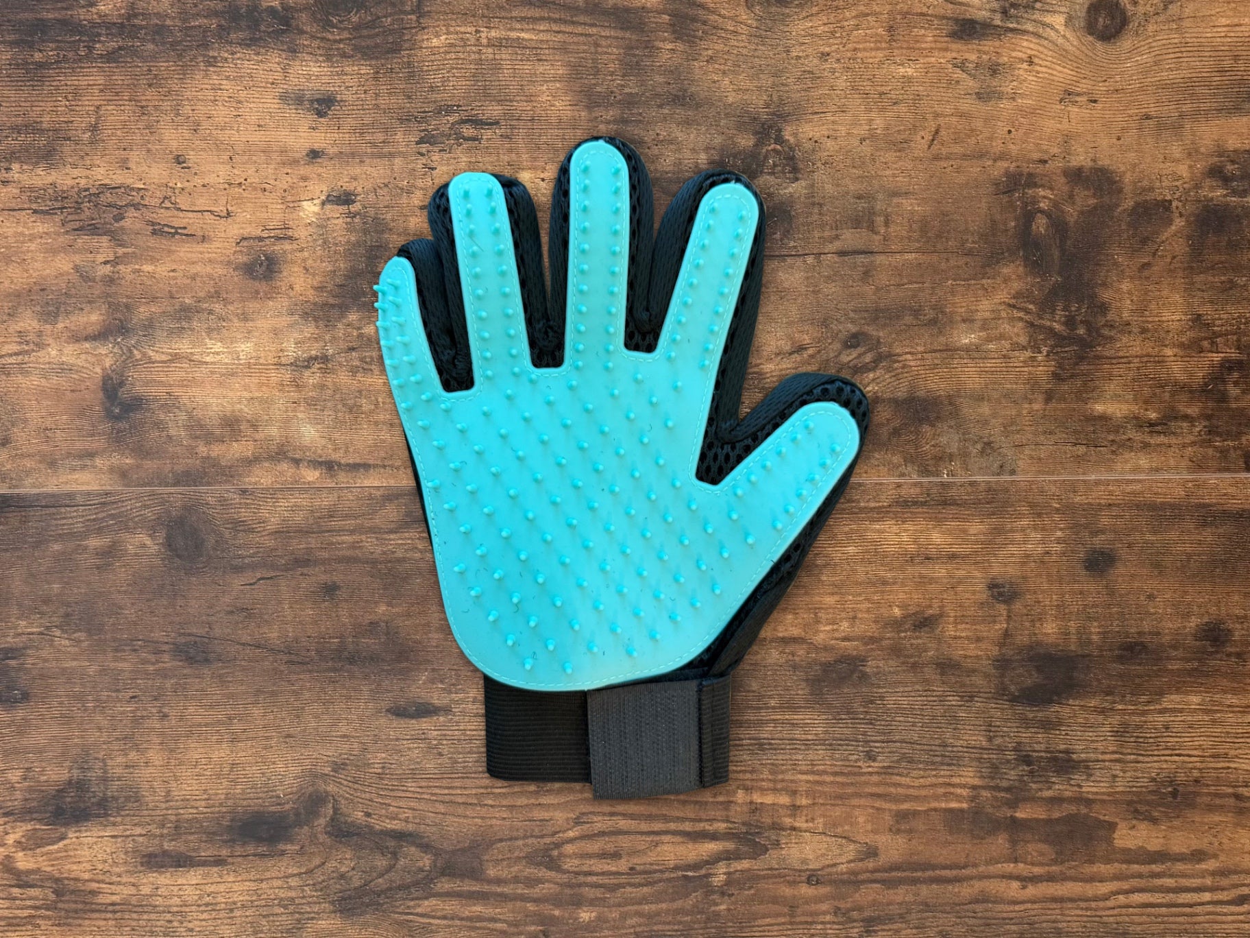 A pet grooming glove with a bright turquoise blue silicone palm and black textured sides, laid flat on a rustic brown wooden surface.
