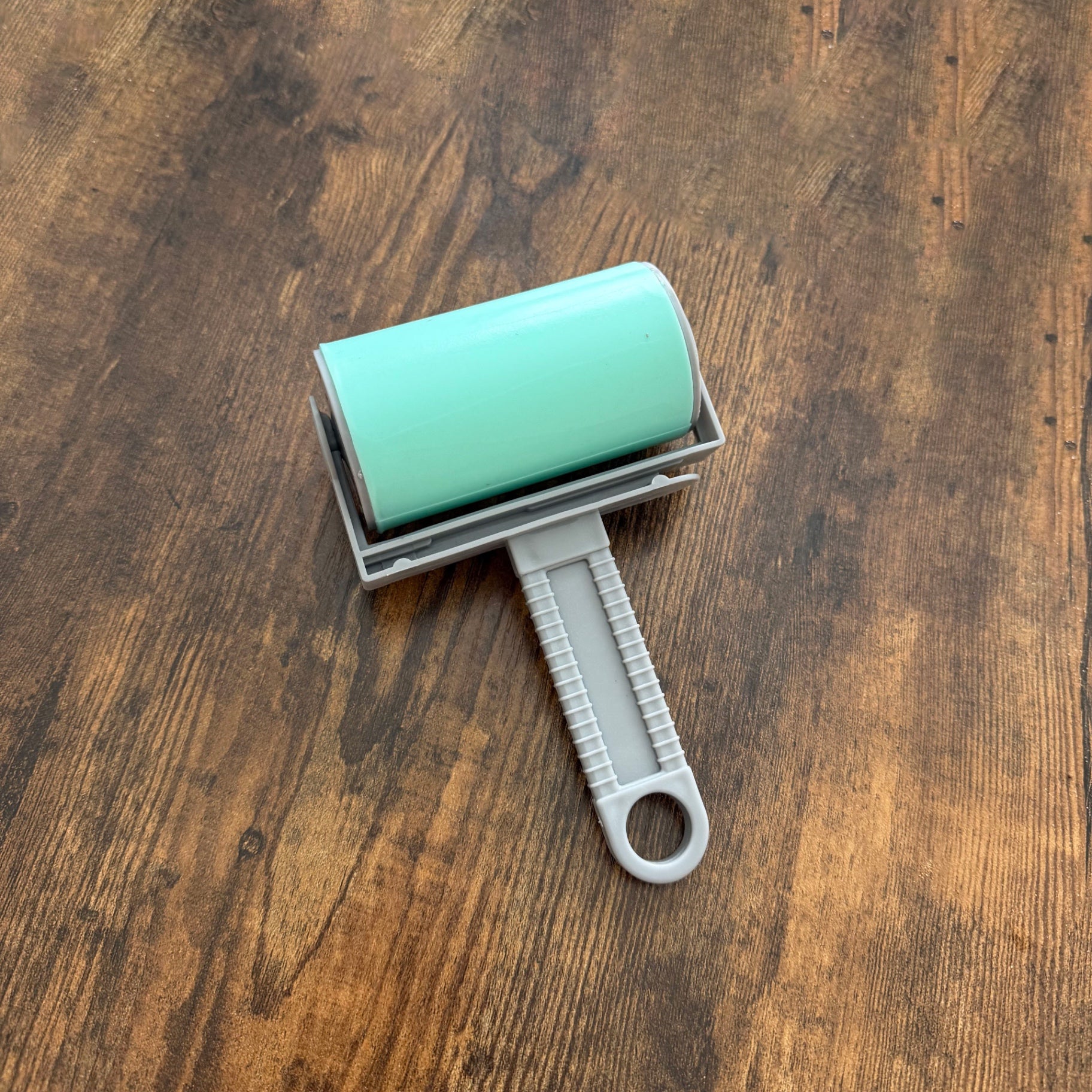 A lint roller with a mint green cylindrical body and a gray handle, shown from a top-down view on a dark brown wooden surface.