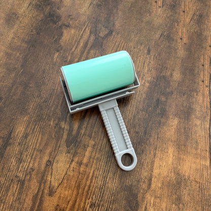 A lint roller with a mint green cylindrical body and a gray handle, shown from a top-down view on a dark brown wooden surface.